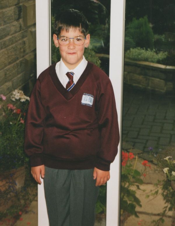 James aged ~7-9 in a loose red school jumper, standing in front of a window and plants with coloured glasses on.
